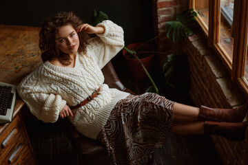 Indoor full-length fashion portrait of young beautiful confident woman wearing cozy white knitted sweater, leather belt, animal print skirt, brown cowboy style boots, posing at loft interior
