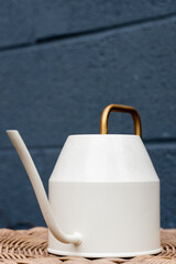 Angled view of a white watering can with a gold handle on a wicker table with a blue cinder block background with space on top