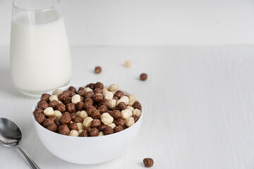 Bowl full of ready-to-eat healthy vegetarian cereals in shape of cocoa and vanilla balls served with spoon and glass of milk prepared for breakfast on white wooden background. Image with copy space