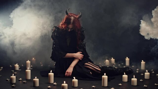 Horned witch spelling, conducting ritual and summon supernatural powers. Candles placing around female demon, smoke rising in the dark. Cult of devil, halloween party or halloween costume concept