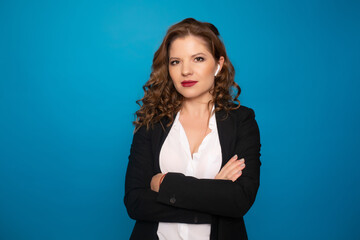 Confident woman with earphones in formal suit covering stomach with crossed arms over blue background. Business concept.