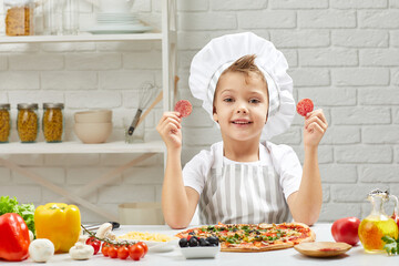 little boy in chef hat and an apron cooking pizza in the kitchen. the child holding sausage slices. having fun