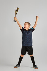 Football player holding a golden cup. little child boy playing soccer and holding trophy on gray...