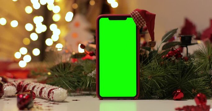 iPhone Xs max in Vertical orientation with green screen on a background of Christmas tree and holiday lights. Santa Claus hat on top. Online store, App promo, ads. Slider Shot