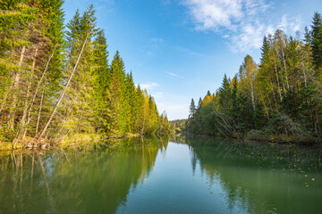 Autumn forest trees are reflected in the river water of the panoramic landscape. Blue sky with clouds.