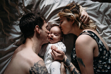 Dad and mom are kissing a emotional little child on the cheek on the bed and the child is laughing