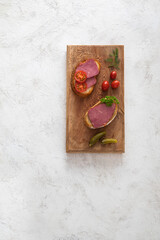 Beef ham sandwiches on wooden cutting board with pickles and greens on white textured background.