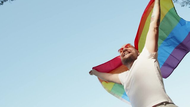 Queer man holding rainbow gay flag while parade on background of blue sky. Happy guy wearing heart sunglasses demonstrate his rights. LGBTQI, Pride Event, LGBT Pride Month, Gay Pride Symbol