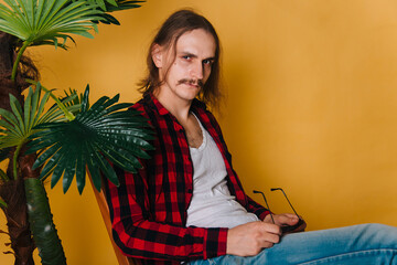 An attractive man with long hair and a mustache in a red plaid 80s disco shirt sits on a chair...