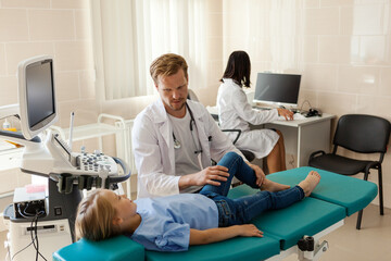 Middle aged male doctor in white coat bending leg of little girl lying on couch to check her knee, female nurse working at desk in background