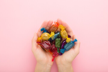 Colorful wrapped chocolate candies in young adult woman palms on light pink table background....