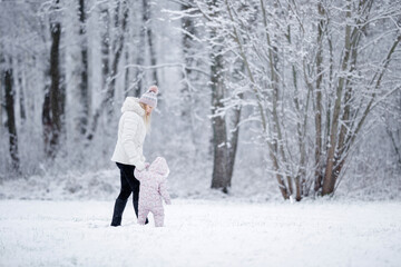 Fototapeta na wymiar Little daughter in overalls and young adult mother walking on white first snow at park. Spending time together in beautiful winter day. Enjoying peaceful stroll.