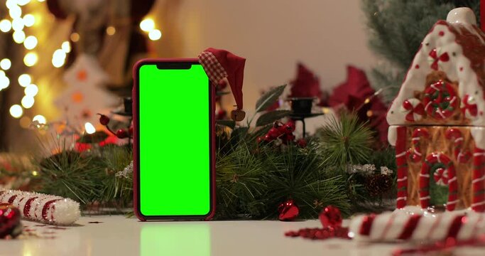 iPhone Xs max in Vertical orientation with green screen on a background of Christmas tree and holiday lights. Santa Claus' hat on top. Online store, App promo, ads.