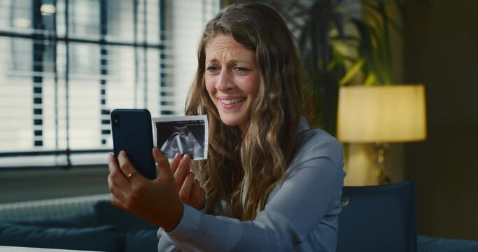 Cinematic slow motion close up shot of an young pregnant woman is making a video call to her relatives or friends and showing an ultrasound to announce her future child birth.