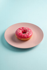 Pink donut with sprinkles on pink plate, sweet glazed dessert food on blue minimal background, angle view