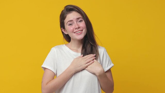Excited smiling pleasant brunette young woman 20s years old in basic white t-shirt posing isolated on yellow background studio. People lifestyle concept. Looking camera holding hands folded on heart