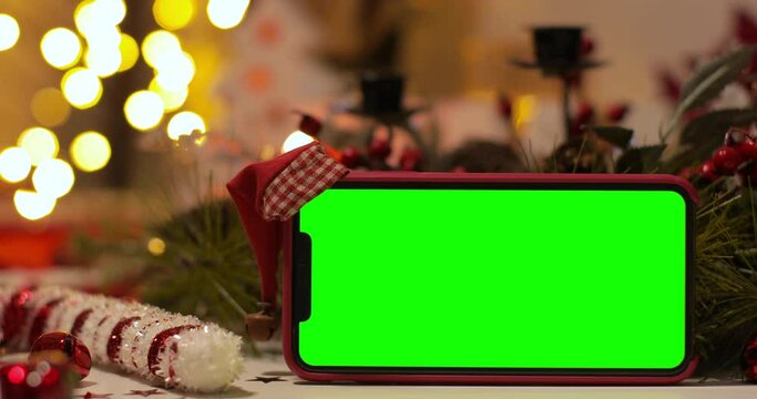 iPhone Xs max in Horizontal orientation with green screen on a background of Christmas tree and holiday lights. Santa Claus' hat on top. Online store, App promo, ads.