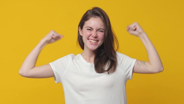 Strong laughing brunette young woman 20s years old wearing casual white t-shirt posing isolated on yellow color background in studio. People lifestyle concept. Looking camera showing biceps muscles