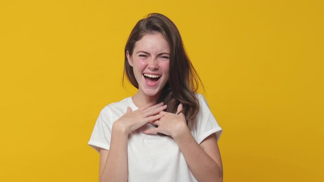 Excited laughing brunette young woman 20s years old in white t-shirt posing isolated on yellow color background in studio. People lifestyle concept. Pointing index finger on camera put hands on chest