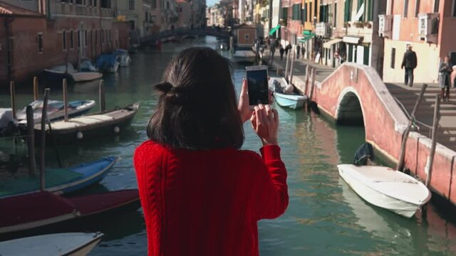 Rear view of beautiful asian girl in red sweater taking photo of the picturesque venetian canal with her smartphone from edge of the bridge. Charming woman spending her vacation in Venice.