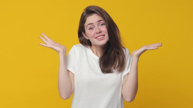 Confused puzzled brunette young woman 20s years old wearing casual white t-shirt posing isolated on yellow color background in studio. People lifestyle concept. Looking camera spreading hands say oops