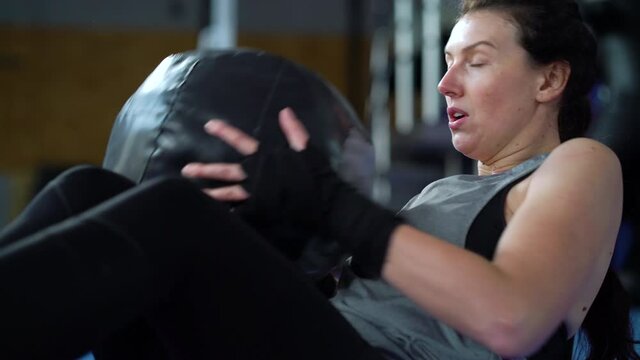 Fit woman in sportswear with bandages on wrists sitting on floor with raised feet and doing crunches with heavy ball. Tracking shot of female boxer training hard in gym. Concept of sport