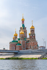 Fototapeta na wymiar large red brick Christian Church with Golden domes on the riverbank under a blue sky