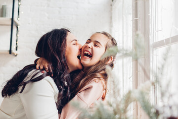 Mother and daughter laughing on kitchen window sill by decorated Christmas tree. Family having fun together at home