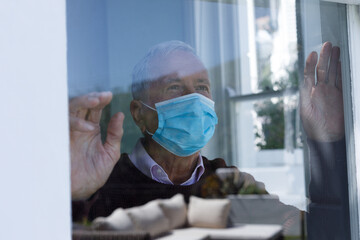 Senior caucasian man wearing face mask looking through glass window at home
