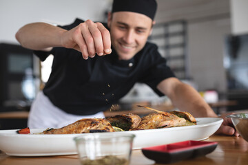 Male caucasian chef adding salt over roasted fish dish in food plate at restaurant kitchen