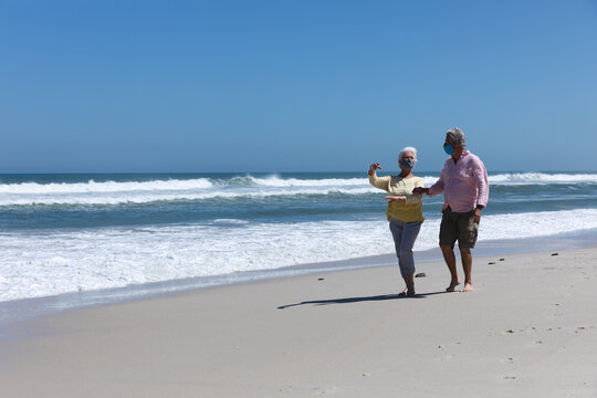 Senior caucasian couple wearing face masks walking on beach talking