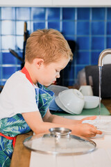 a boy in an apron washes dishes in the kitchen at home.