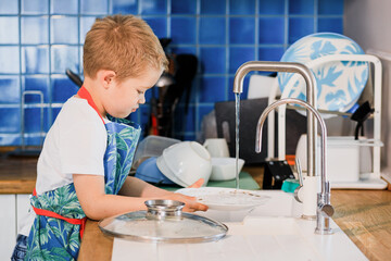 a boy in an apron washes dishes in the kitchen at home.