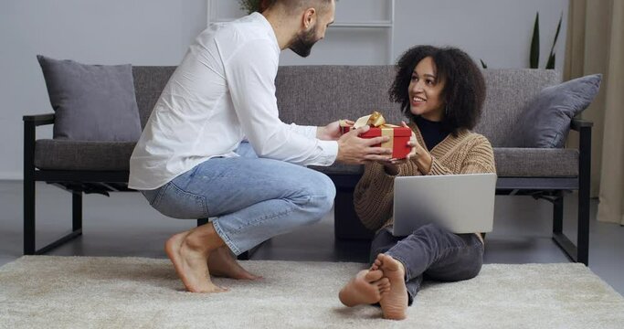 Boyfriend gives girlfriend present in living room. Young african black business woman student sitting with laptop on floor receives red gift box from her caucasian husband, girl feels delight surprise