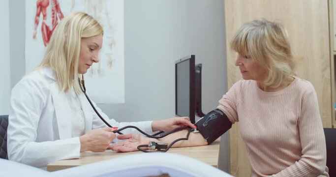 Side view. Portrait of Caucasian beautiful female doctor in white coat sitting in clinic cabinet measuring blood pressure of senior woman patient with medical manometer at appointment. Health concept