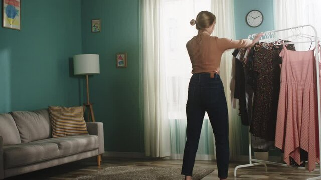 Young girl in jeans and brown sweater in front of window removes dress from hanger and scatters it around spacious room