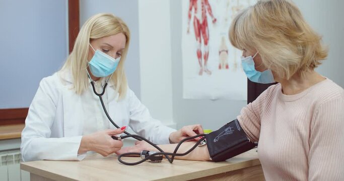 Portrait of Caucasian female doctor in medical mask sitting in clinic and measuring blood pressure of senior woman patient with manometer. Healthcare. Coronavirus pandemic concept