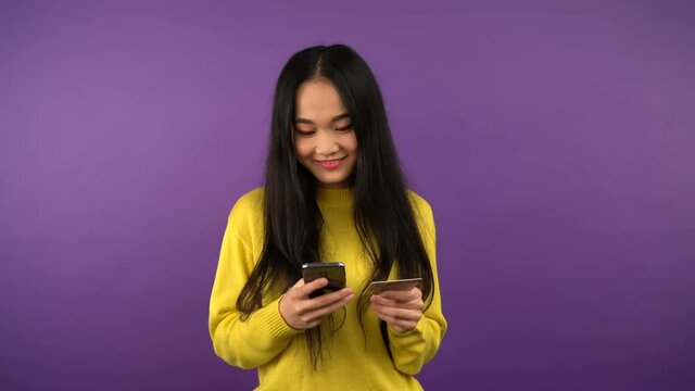 smiling young asian woman in bright yellow sweater is shopping online and paying with gold card via smartphone. 4K