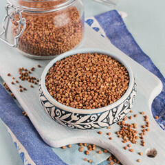 Bowl of dry raw buckwheat groats and glass jar on a light blue background. Cooking buckwheat porridge concept.