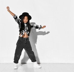 Portrait of an Asian girl with black hair who dances and has fun on a white background. The child wears a black hat, plaid shirt, jeans and looks into the camera. Children's fashion and style.