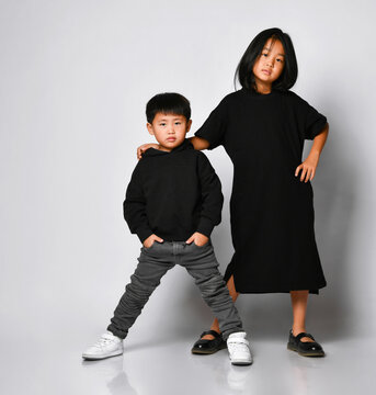 Stylish Brother And Sister Asian Children Posing In Front Of The Camera In The Studio On A White Background. Cool Boy In Black Hoodie And Jeans With Older Sister In Black Dress Having Fun.