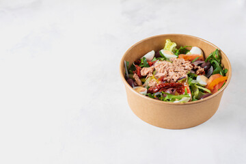 Fish diet salad with tuna, tomatoes, arugula on light background top view. Healthy lunch. Eco-friendly carton packaging, environment protection. Food delivery in disposable plate of craft paper.