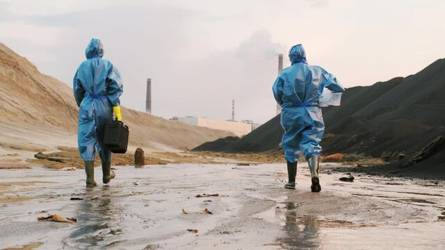 Back-view shot of two ecologists in protective suits and rubber boots walking down muddy area of polluted town outskirts holding equipment for quality control