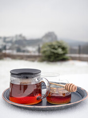 Glass kettle of hot tea and jar of honey on table covered by snow. Landscape on blurry background. Close up with copy space.