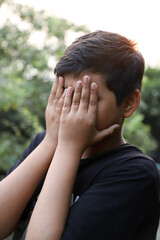 A southeast Asian brown young boy with short hair hiding or covering his face with his two hands-a conceptual vertical photo