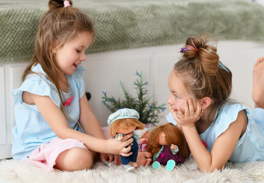 Cheerful pretty small girls sisters kids with long hair in same clorhing playing with toys dolls together having fun at home. Happy childhood, cheerful lifestyle, games, comfortable pastime at home
