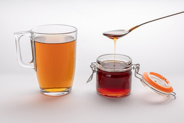 A cup of invigorating tea with spoon of honey, glass cup with drink and honey jar on white background, golden honey spilling from the spoon