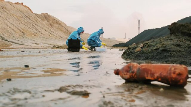 Full shot of ecology specialists in blue protective costumes squatting getting puddle water to test tube for quality control test examining water pollution in dirty industrial town