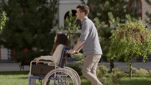 Young man riding his disabled invalid girlfriend on wheelchair talking and laughing. Asian handicapped girl with paralysis living full life. Couple in love. 6k downscale 10 bit color.