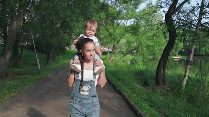 Cute little boy sitting on mom's shoulders discovering the world around. Beautiful mother and her sweet son walking in the park near picturesque pond. Concept of motherhood, happiness , love.